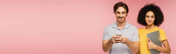 Joven mujer hispana con el ordenador portátil sonriendo a la cámara cerca de hombre charlando en el teléfono inteligente aislado en rosa, bandera - foto de stock