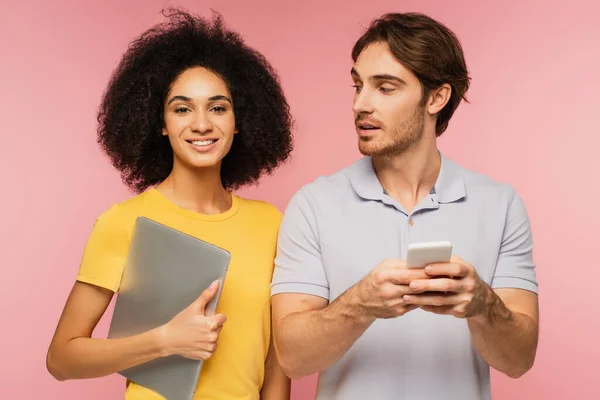 Heureuse femme hispanique avec ordinateur portable regardant la caméra près de l'homme en utilisant un téléphone cellulaire isolé sur rose — Photo de stock