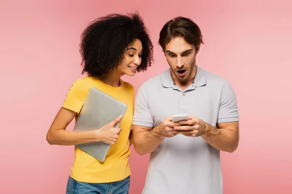 Uomo stupito utilizzando il telefono cellulare vicino sorridente donna ispanica in possesso di laptop isolato su rosa — Foto stock