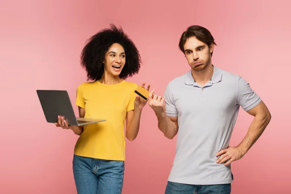 Unzufriedener Mann bläst Wangen, während er seiner hispanischen Freundin mit Laptop Kreditkarte gibt, isoliert auf rosa — Stockfoto