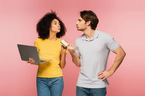 Feliz pareja interracial con portátil y tarjeta de crédito soplando aire beso el uno al otro aislado en rosa - foto de stock