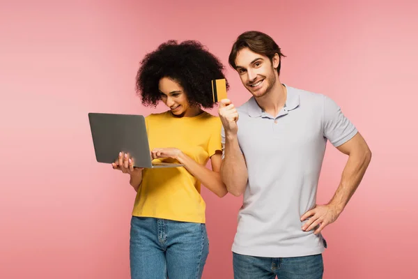 Hombre feliz mostrando tarjeta de crédito mientras está de pie con la mano en la cadera cerca de la mujer latina utilizando el ordenador portátil aislado en rosa - foto de stock