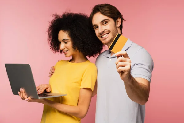 Souriant hispanique femme à l'aide d'un ordinateur portable près homme joyeux tenant carte de crédit isolé sur rose — Photo de stock