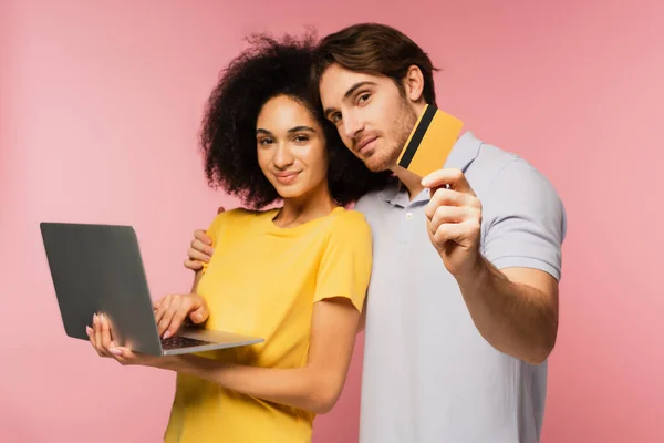 Чоловік показує кредитну картку і обіймає свою іспанську дівчину за допомогою ноутбука ізольовано на рожевому — стокове фото