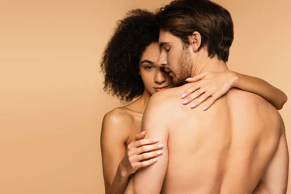 Seductive latin woman looking at camera while hugging sexy shirtless man isolated on beige — Stock Photo