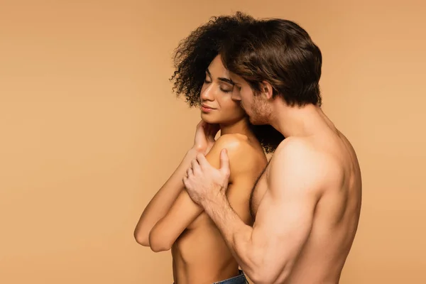 Joven hombre sin camisa abrazando topless mujer hispana de pie con los ojos cerrados aislados en beige - foto de stock