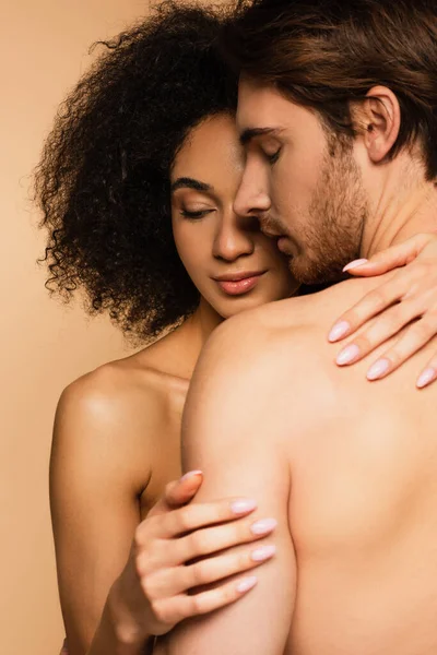 Mujer latina joven y en topless abrazando al hombre sin camisa aislado en beige - foto de stock