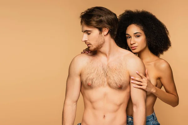 Topless latin woman looking at camera while embracing sexy shirtless man isolated on beige — Stock Photo