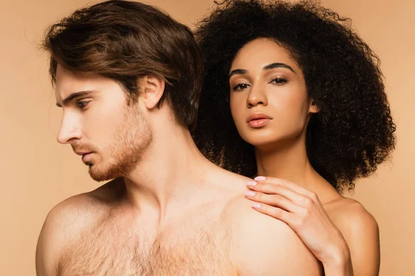 Seductora mujer hispana abrazando hombro de hombre sin camisa mientras mira la cámara aislada en beige — Stock Photo