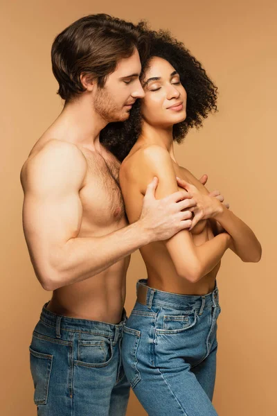 Без сорочки чоловік в джинсах обіймає сексуальну латинську жінку, ховаючись грудьми з схрещеними руками ізольовані на бежевому — стокове фото