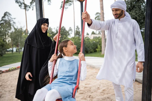 Smiling muslim parents standing near daughter on swing outdoors — Stock Photo