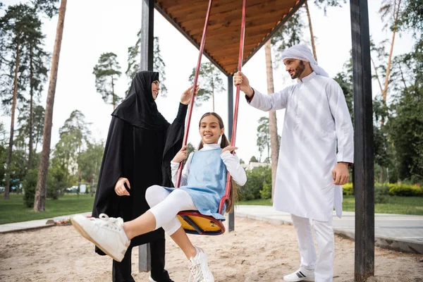 Smiling arabian child on swing near parents in park — Stock Photo