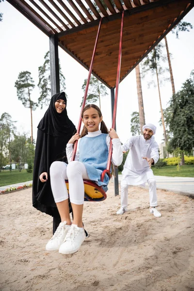 Joyeux arabian fille balançant sur aire de jeux près des parents dans le parc — Photo de stock