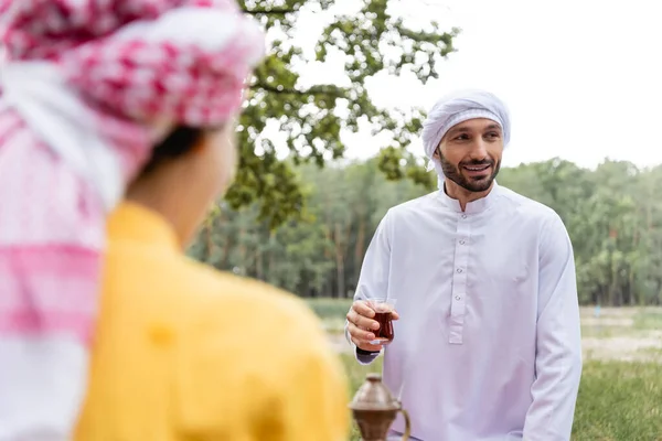 Smiling muslim man holding glass of tea near blurred son outdoors — Stock Photo