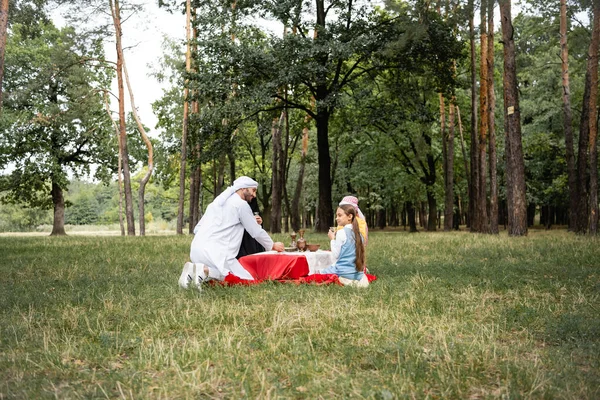 Padre árabe positivo sentado cerca de la familia durante el picnic en el parque - foto de stock