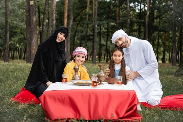 Arabian family smiling at camera near food and tea in park — Stock Photo