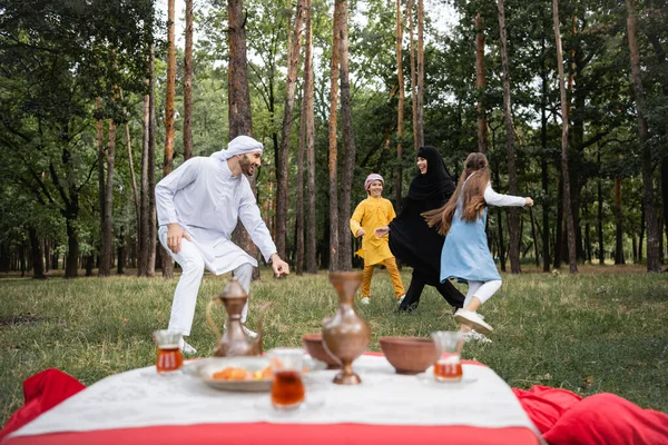 Positive muslim family in traditional clothes playing near tea on blurred foreground in park — Stock Photo