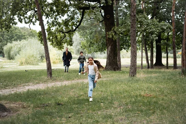 Menina muçulmana correndo no gramado perto do irmão e dos pais no parque — Fotografia de Stock