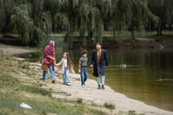 Familia árabe positiva cogida de la mano mientras camina cerca del lago - foto de stock
