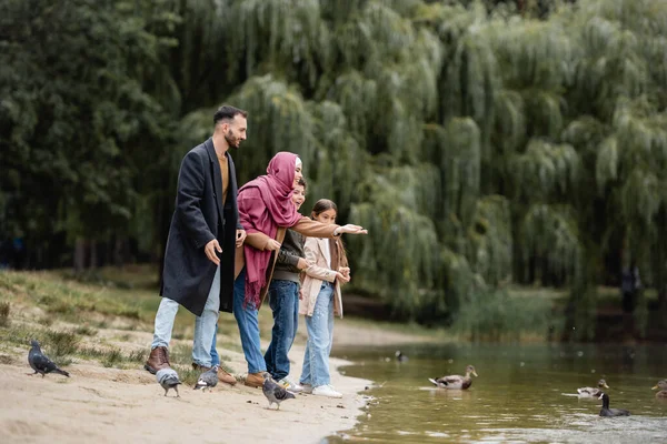 Sonriente madre musulmana señalando a los pájaros en el lago cerca de marido e hijos en la playa - foto de stock