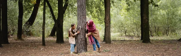 Sonriente chica árabe jugando con la madre cerca del árbol en el parque, pancarta - foto de stock