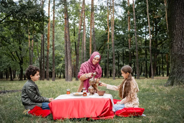 Усміхнена мусульманська мати виливає чай біля дітей у парк — стокове фото