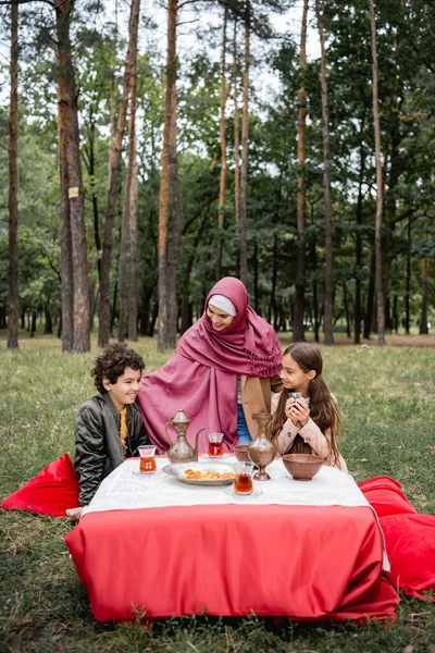 Positive arabische Mutter umarmt Kinder in der Nähe von Tee und Trockenfrüchten im Park — Stockfoto