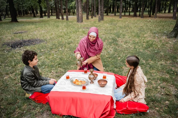 Donna musulmana versando tè vicino ai bambini e frutta secca nel parco — Foto stock