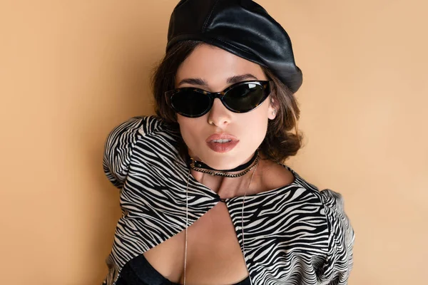 Top view of brunette woman in zebra print outfit, leather beret and sunglasses looking at camera on beige — Stock Photo