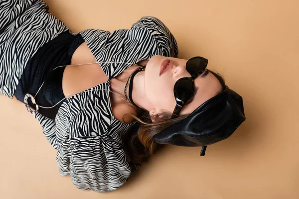 Vista de ángulo alto de la mujer joven en traje de impresión de cebra, boina de cuero y gafas de sol posando en beige - foto de stock