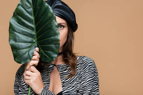 Mujer joven en traje de impresión de cebra y boina de cuero con hoja verde mientras cubre la cara aislada en beige - foto de stock