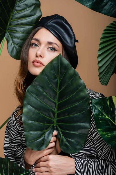 Jovem mulher em boina de couro perto de folhas tropicais verdes isoladas em bege — Fotografia de Stock