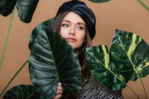 Junge Frau mit Lederbarett in der Nähe tropischer Blätter isoliert auf beige — Stockfoto