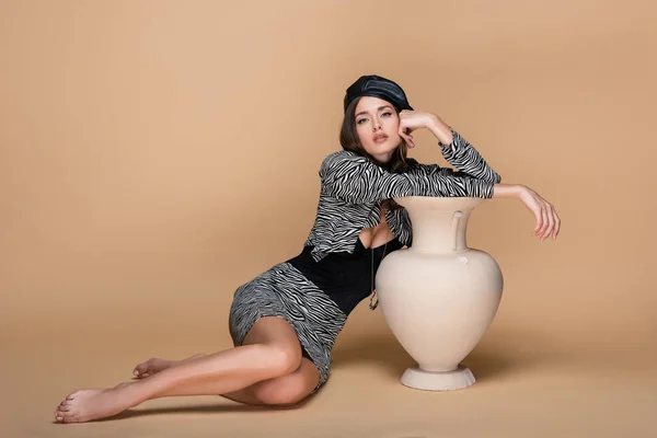 Barefoot woman in zebra print outfit and black beret posing near clay vase on beige — Stock Photo