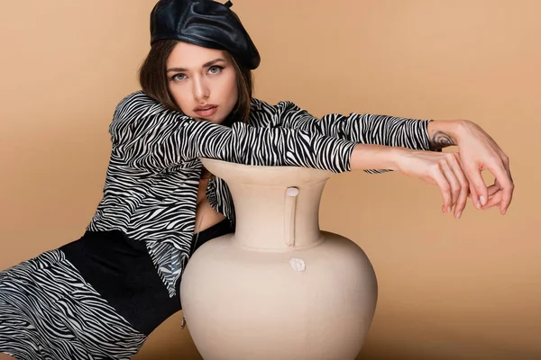 Young tattooed model in zebra print outfit and black beret posing near clay vase on beige — Stock Photo