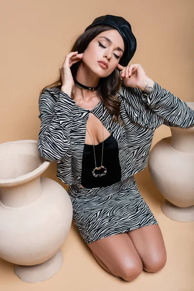 Hübsches Model im Zebra-Print-Outfit und schwarzer Ledermütze posiert in der Nähe von Tonvasen auf Beige — Stockfoto