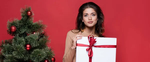 Hübsche Frau mit eingewickeltem Geschenk in der Nähe des Weihnachtsbaums auf rotem Banner — Stockfoto