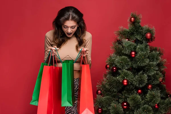 Mujer joven y sorprendida sosteniendo bolsas de compras cerca del árbol de Navidad aislado en rojo - foto de stock