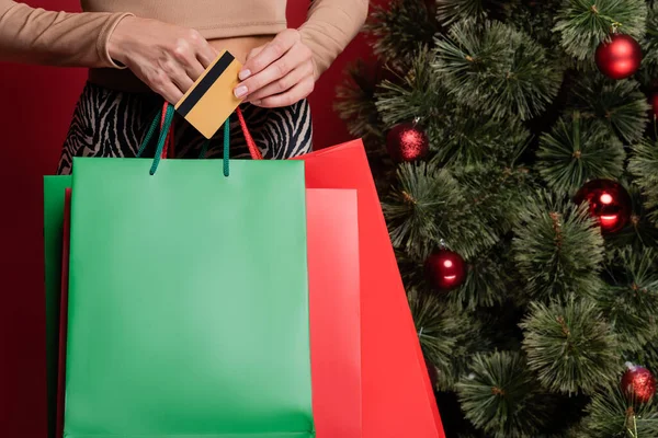 Vista ritagliata di giovane donna che tiene borse della spesa e carta di credito vicino all'albero di Natale isolato sul rosso — Foto stock