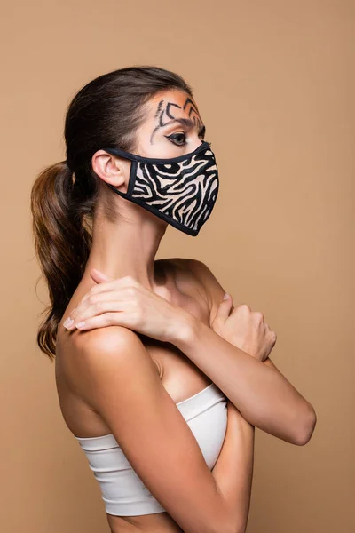 Mujer joven con maquillaje de tigre y máscara protectora de impresión animal posando aislado en beige - foto de stock