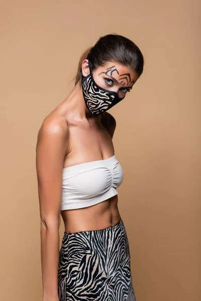 Брюнетка с макияжем тигра и защитной маской для печати животных, позирующей изолированно на бежевом — стоковое фото
