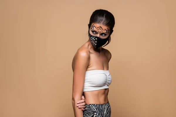 Woman with tiger makeup and animal print protective mask looking away isolated on beige — Stock Photo