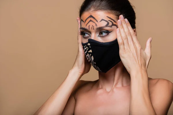 Modelo con maquillaje de tigre y máscara protectora animal print mirando hacia otro lado aislado en beige - foto de stock