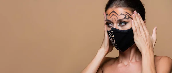 Modelo con maquillaje de tigre y máscara protectora de impresión animal mirando hacia otro lado aislado en beige, bandera - foto de stock