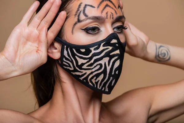 Modelo joven con maquillaje de tigre y máscara de animal print mirando hacia otro lado aislado en beige - foto de stock