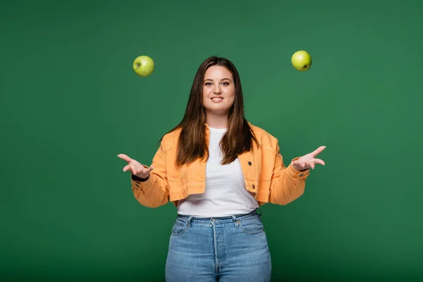 Mujer sonriente con sobrepeso lanzando manzanas aisladas en verde - foto de stock