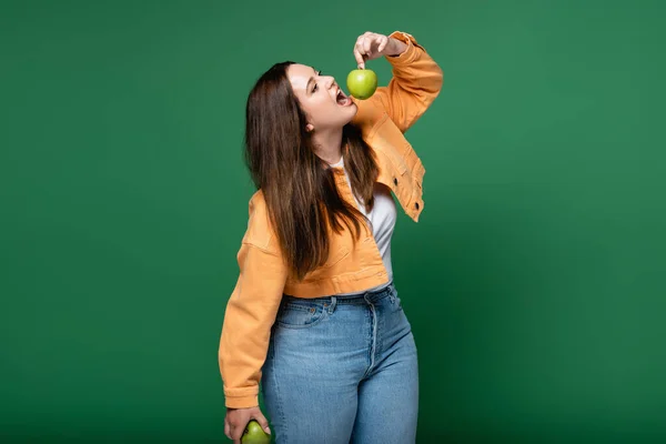 Молода жінка плюс розмір тримає яблуко біля відкритого рота ізольовано на зеленому — стокове фото