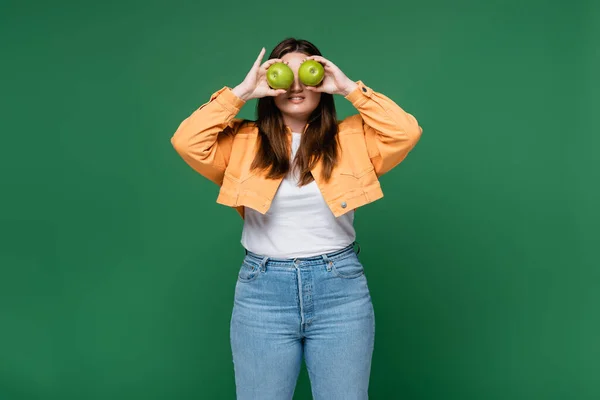 Улыбающаяся женщина с избыточным весом держит зеленые яблоки возле глаз изолированы на зеленый — стоковое фото