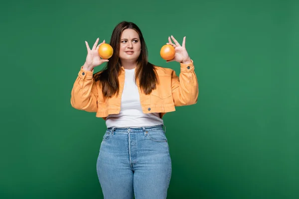 Confused woman with overweight holding oranges isolated on green — Stock Photo