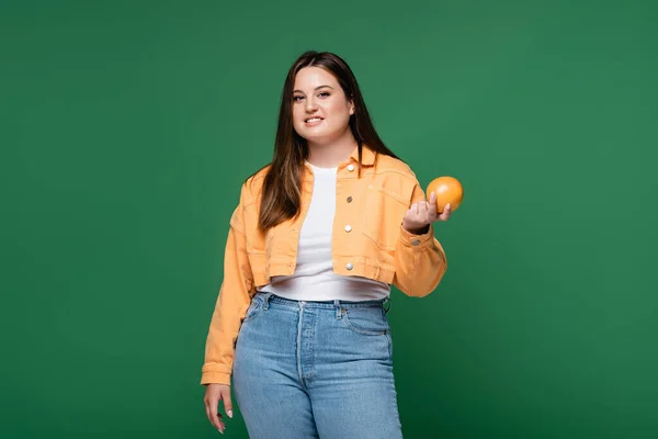 Mujer positiva con sobrepeso sosteniendo naranja y sonriendo a la cámara aislada en verde - foto de stock